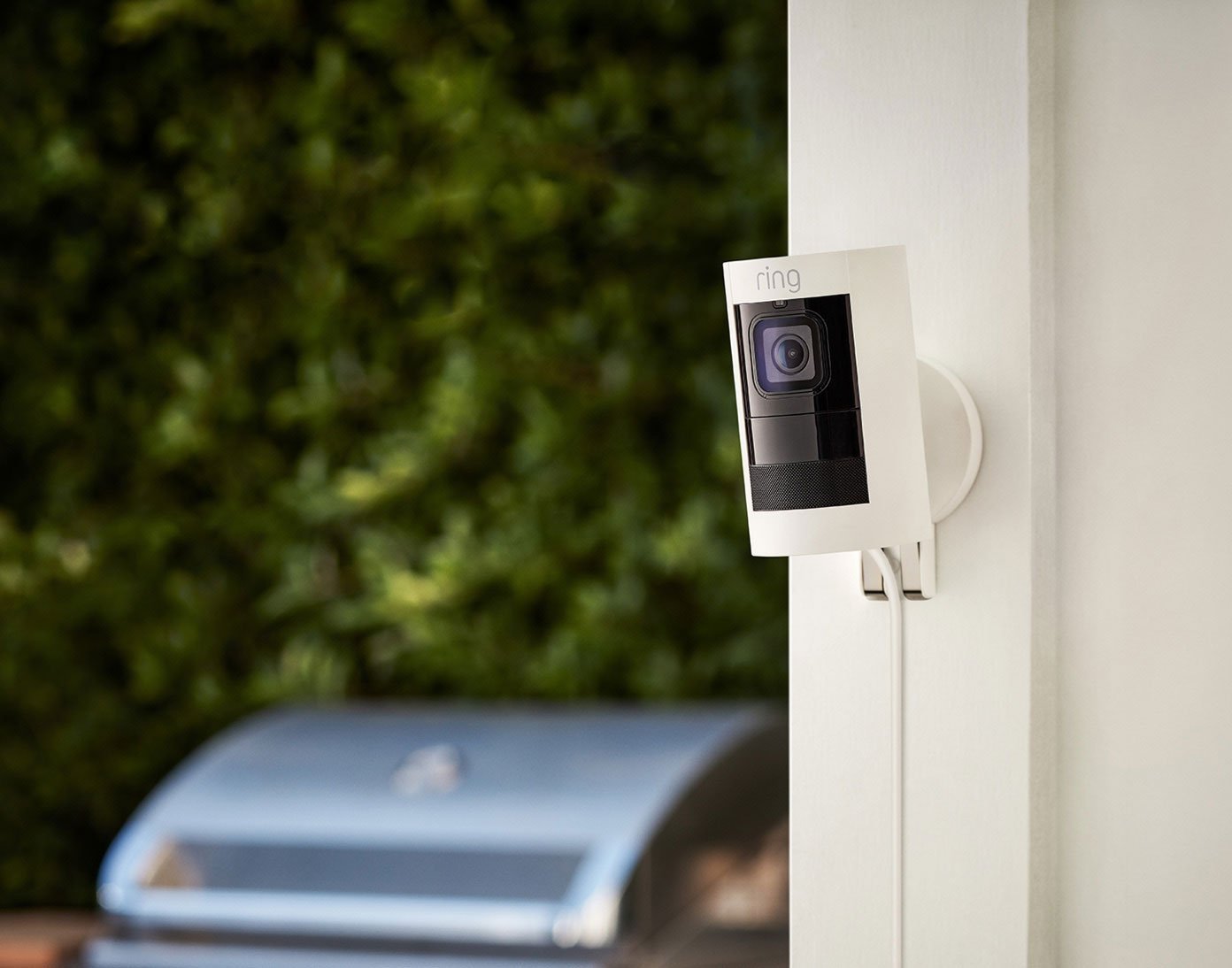 Ring Stick UP Cam Elite mounted on outdoor wall near barbecue grill with green hedge in background.
