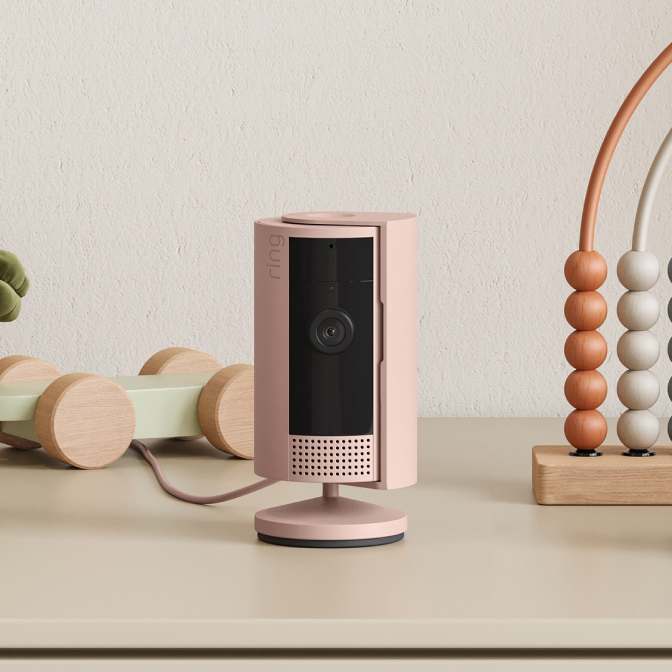 Blush pink Ring Indoor Camera (2nd Gen) on a table between wooden toys, with privacy cover partially open.