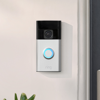 Ring Battery Video Doorbell mounted beside door, with camera lens and blue-lit button on satin-nickel faceplate.