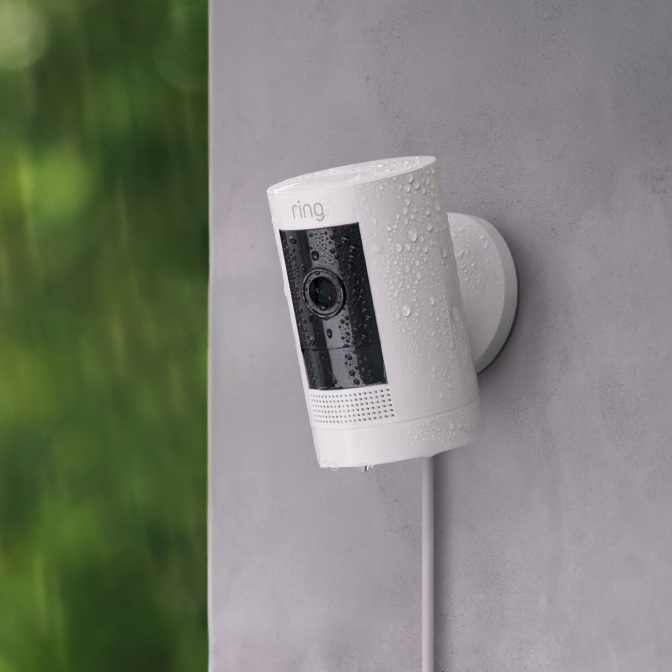 White Stick Up Cam Plug-In with power cord visible, mounted on grey wall outdoors.