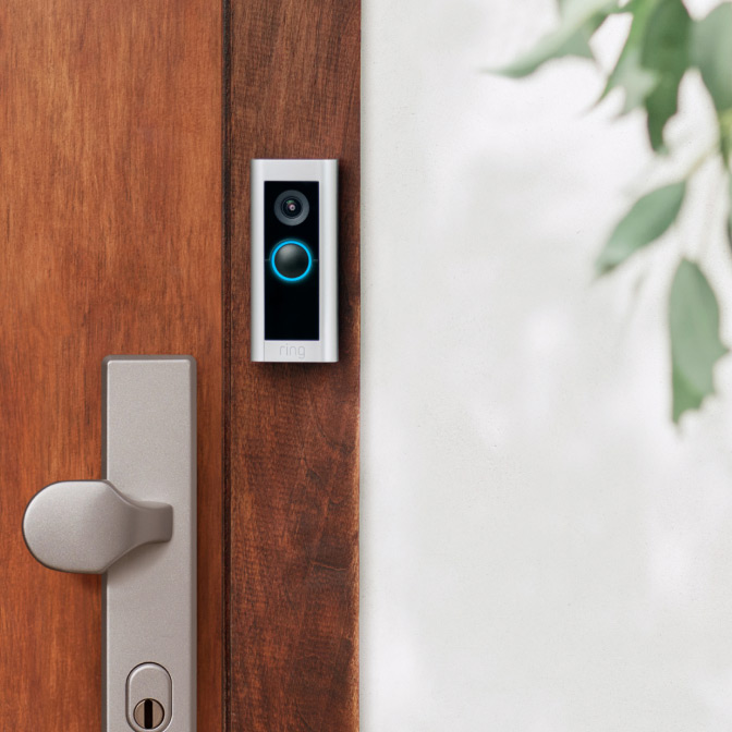 Ring Wired Video Doorbell mounted next to wooden door, with camera lens and blue-lit button on satin-nickel frame.