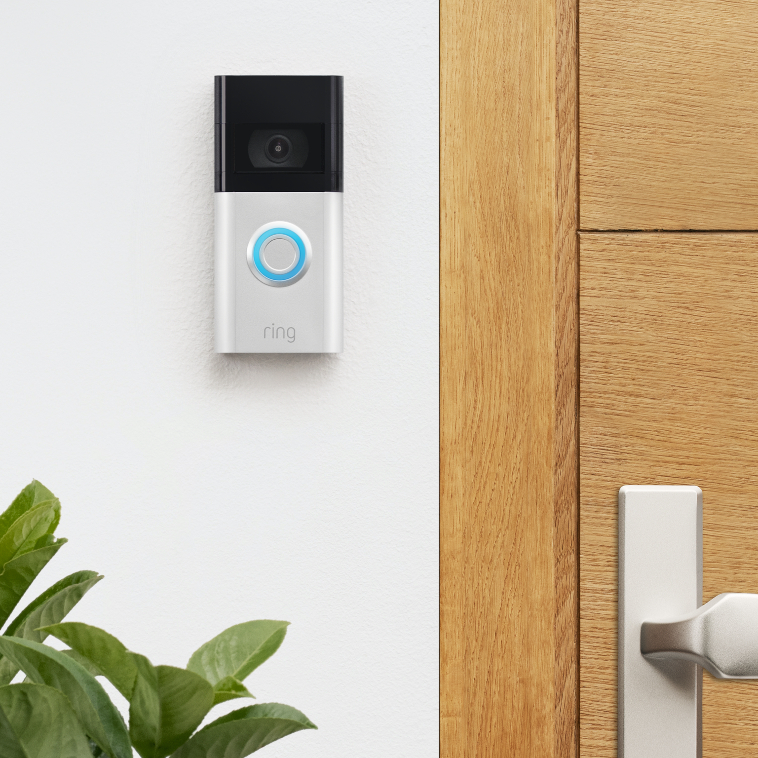 Ring Video Doorbell with blue-lit button mounted on white wall beside a wooden door with silver handle.