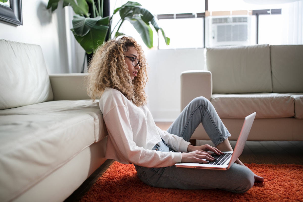Pessoa trabalhando com um laptop no colo sentada em um tapete no chão; ambiente informal de home office