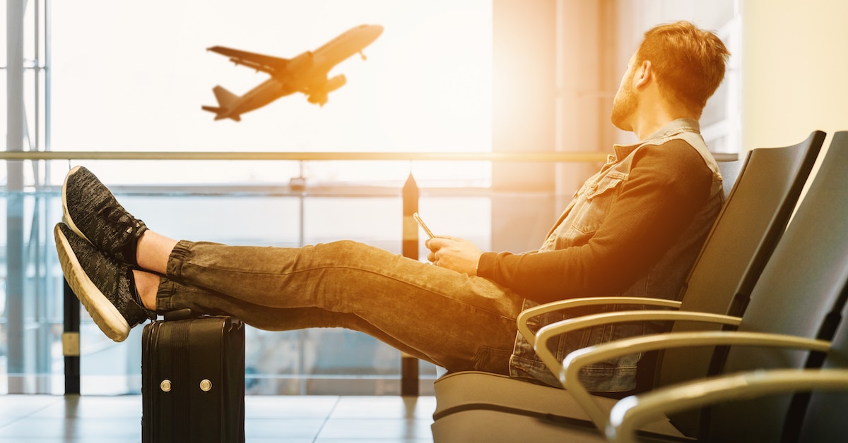 Homem no aeroporto, com os pés em cima da mala, vendo avião decolar