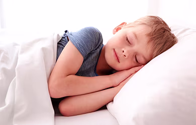 A boy lying on a bed with white bedding wearing a blue striped shirt in a bright bedroom.