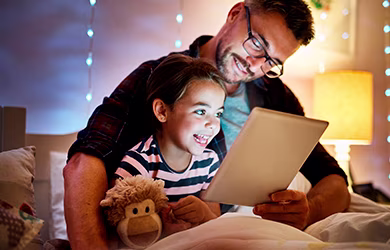 A father and child smiling while watching a tablet in a room with a bed lamp turned on.
