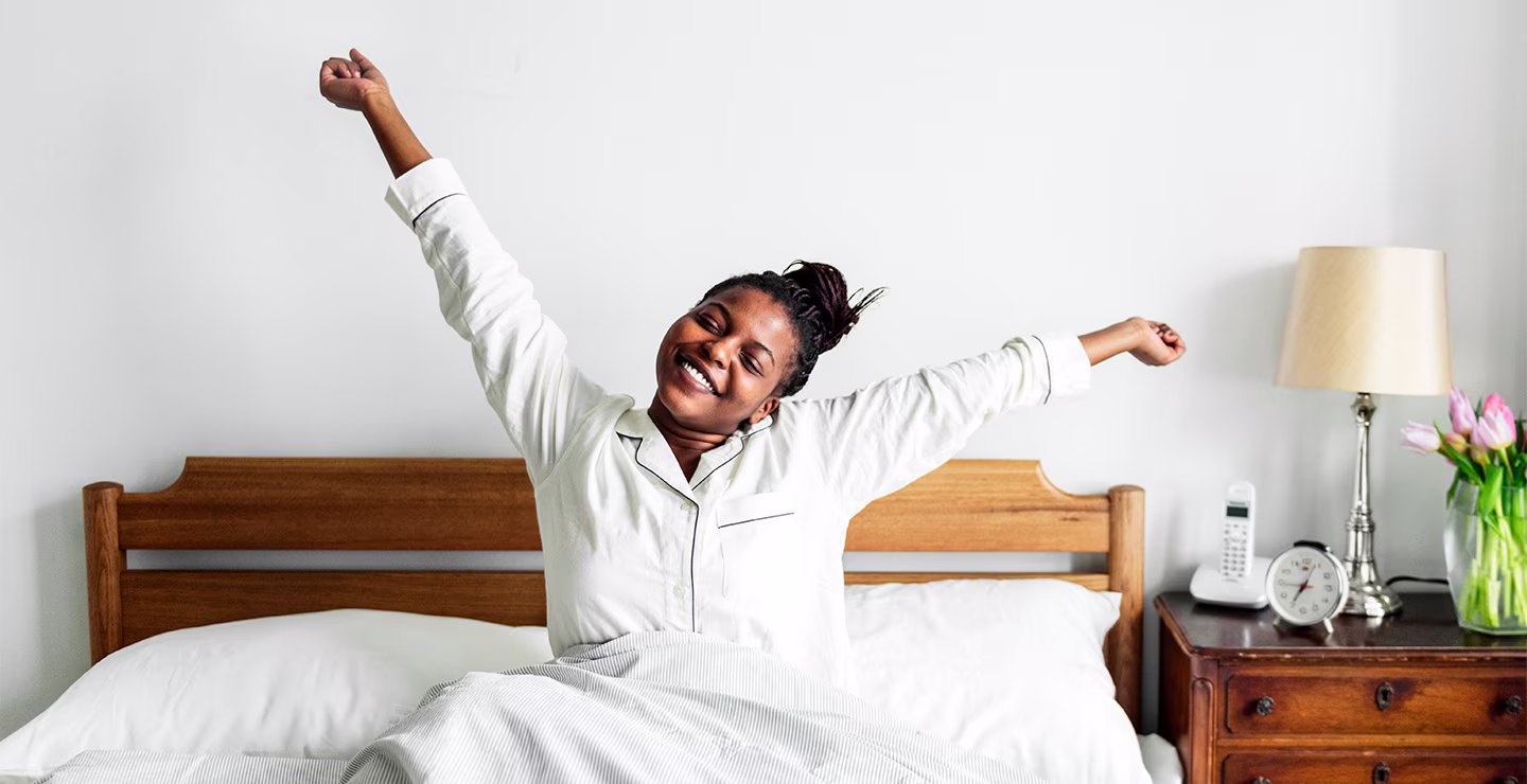 A woman in pajamas stretching arms while sitting on a bed with white bedding and wooden headboard.