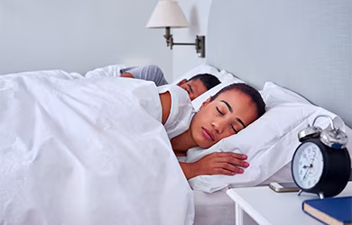 Two people sleeping in a bed with white bedding and a nightstand with clock and book.