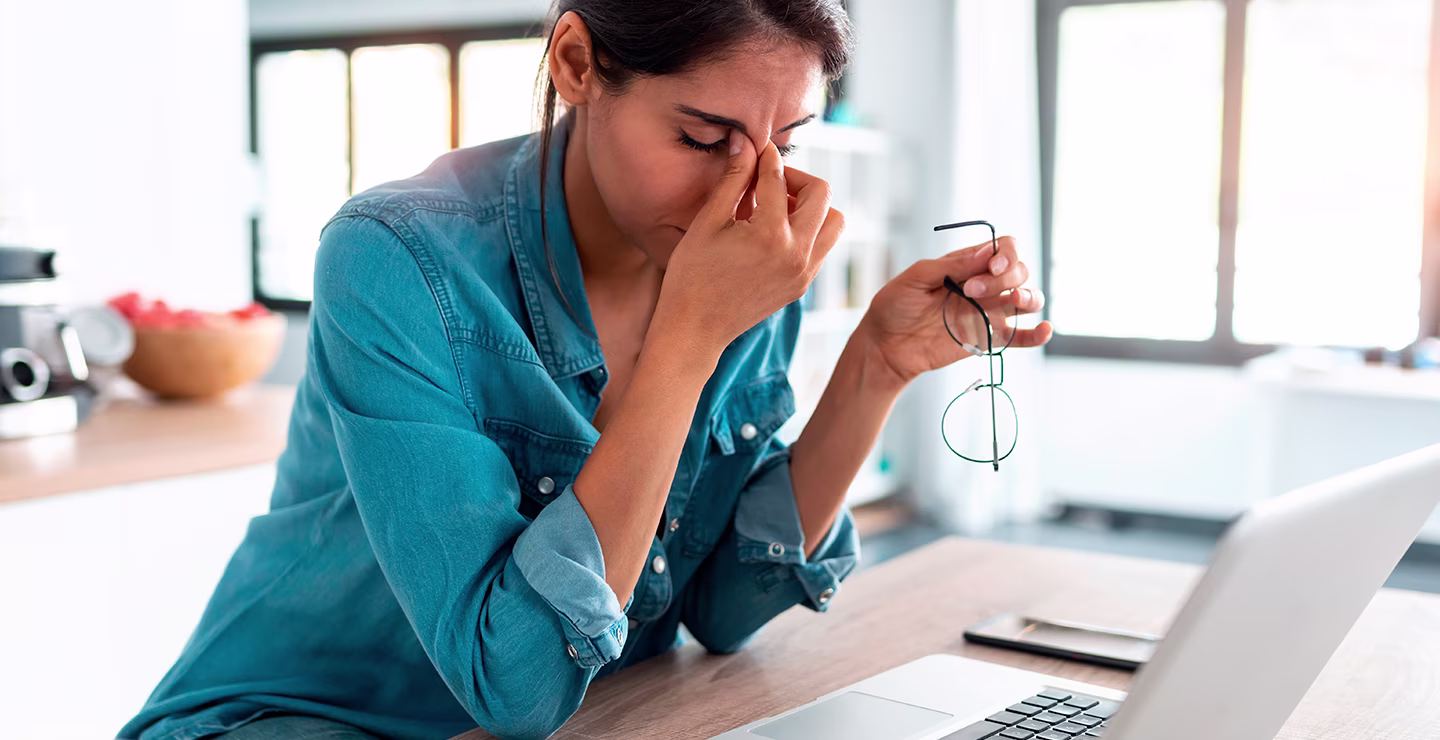 A woman sitting at a table with a laptop, holding her glasses and pinching the bridge of her nose, appearing stressed or experiencing eye strain.