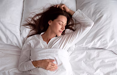A woman with brown hair sleeps peacefully in a white bed, her hand resting near her head.