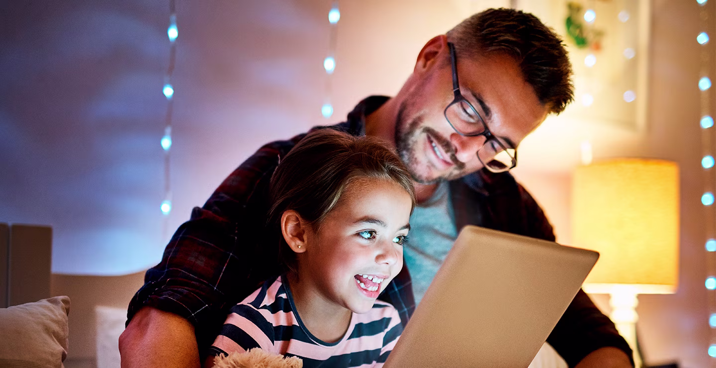 A father and child smiling while watching a tablet in a room with a bed lamp turned on.
