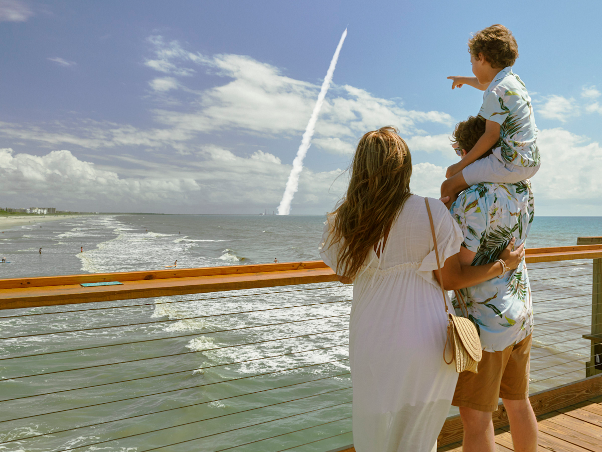 A photo taken from behind of a woman and a man with a child on his shoulders pointing at a rocket taking off in the distance. They are all standing on a pier with the ocean beneath.