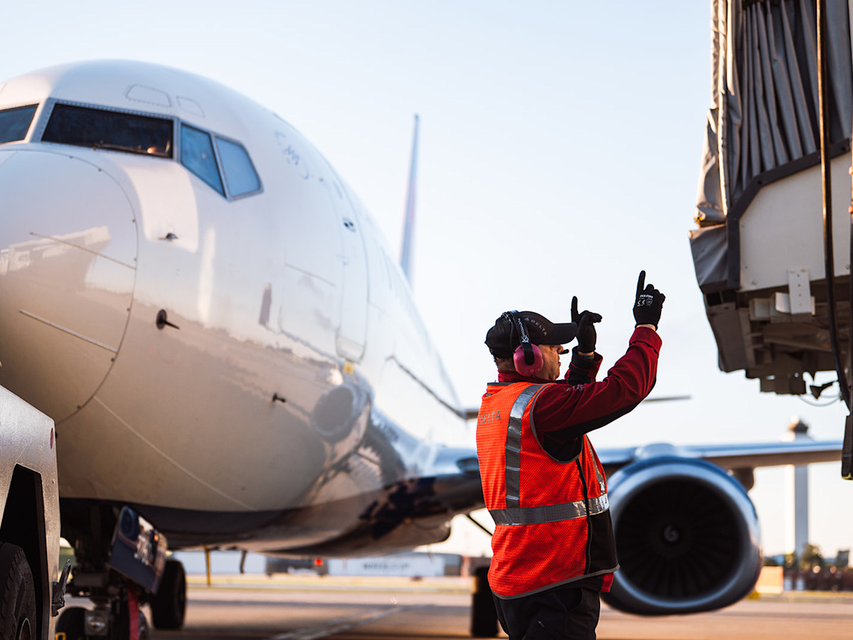 A photo of a man dressed in a safety vest with his hands up to direct plane movement. Behind him is a Delta plane attached to a tug with a disconnected jet bridge to the right.