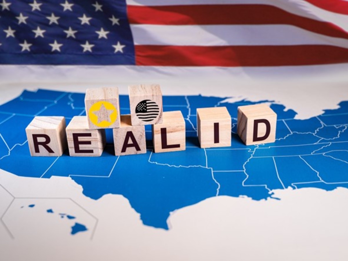 A photo of wooden blocks that spell out Real ID and have a star and an American flag sit on top of a blue map of the United States of America. There is an American flag in the background.