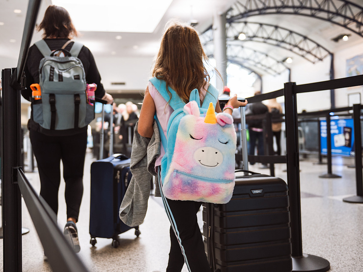 A photo of a little girl in the TSA line with a black suitcase and a unicorn rainbow backpack.