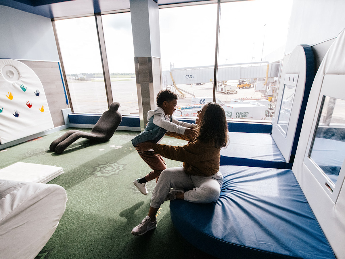 A photo of a happy son running to be embraced by his mom in the Sensory Room in Concourse A. In the Sensory Room is squishy furniture and padding with natural lighting.