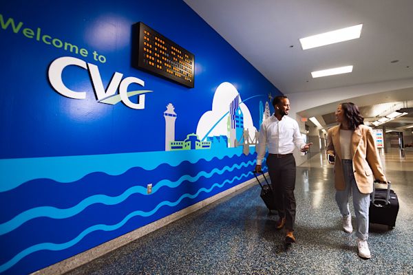 A photo of a young woman and man smiling at one another as they walk with their luggage. To the left is a blue wall that says Welcome to CVG and has an abstract mural of iconic Cincinnati and Northern Kentucky buildings.