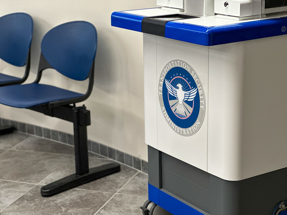 A photo of a cart on wheels with the TSA emblem in the center. There are blue plastic chairs seen in the background.