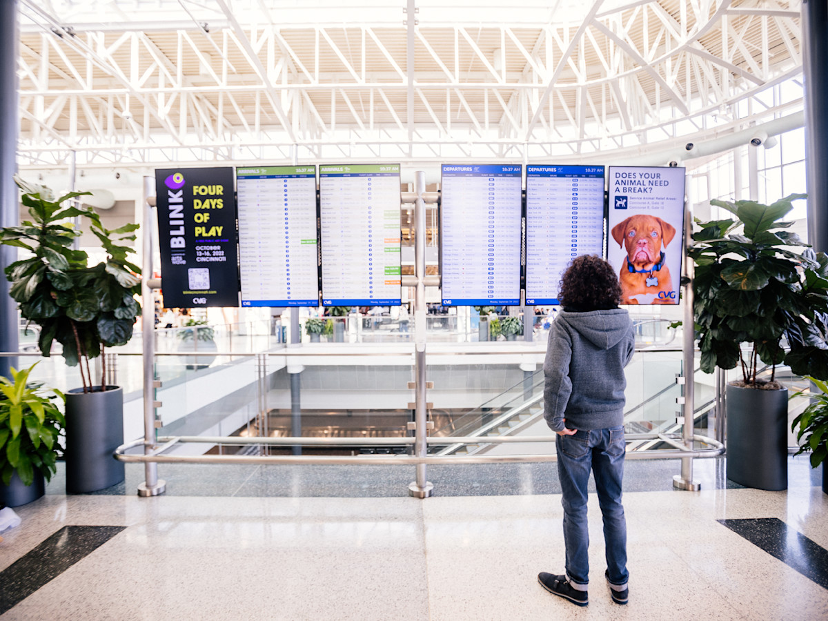 A photo of Jay Kruz standing in front of the Flight Information Display Systems (FIDS) at CVG Airport.