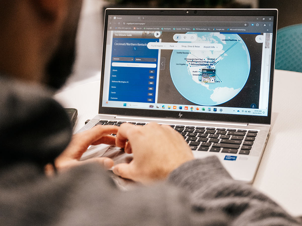 Photo taken from behind a male in a gray sweatshirt with a red baseball cap looking at a laptop with a global map on it.