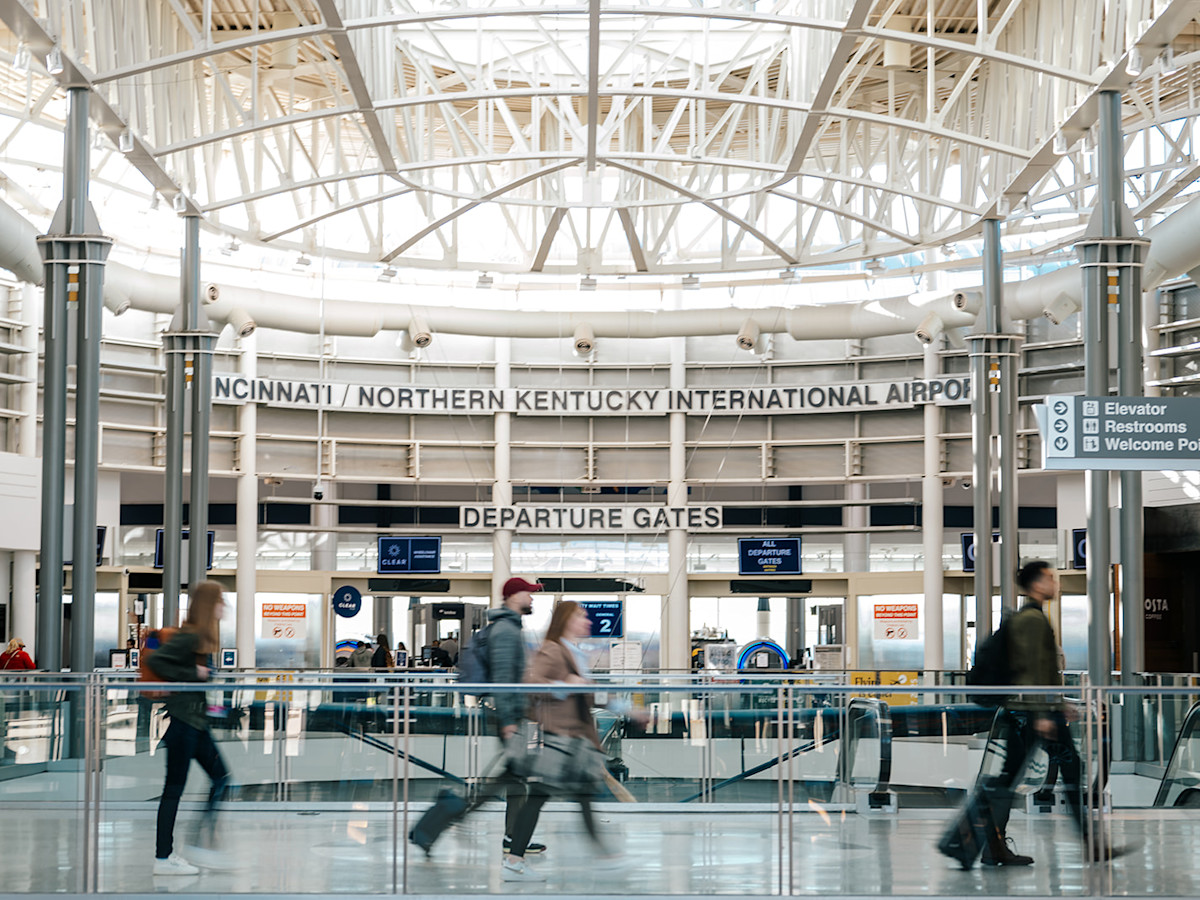 A photo of travelers walking across the Ticketing Bridge at CVG Airport with the TSA checkpoint in the background. The words Cincinnati / Northern Kentucky International Airport and Departure Gates can be seen on the wall in the background.