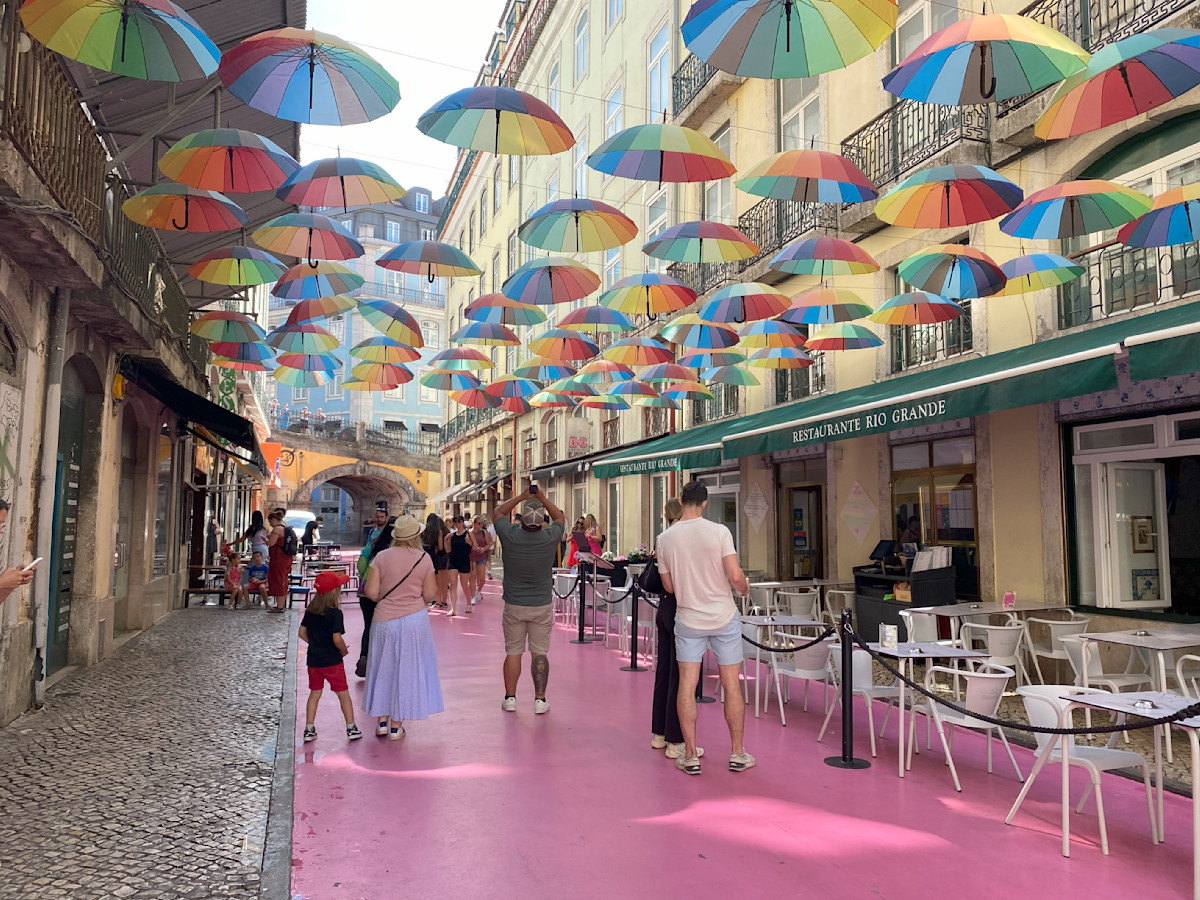 A photo of a pink street with colorful umbrellas suspended above the road. There are cafes on the right with cafe tables and chairs out front. In the distance is a yellow arched bridge. People are walking down the street and taking photos.