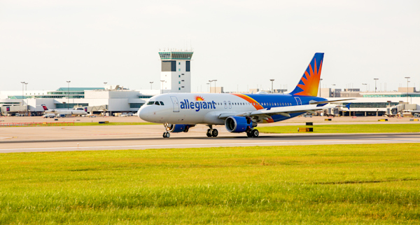 An Allegiant plane landing at CVG. The airport is in the background and in the foreground is bright green grass. It looks like a bright and sunny day.