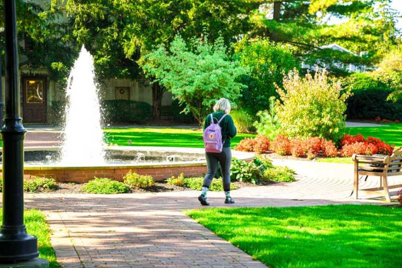 在一个晴朗的夏日里，伊利湖学院校园里基尔考利大厅前的喷泉, with a student walking in front of it. 