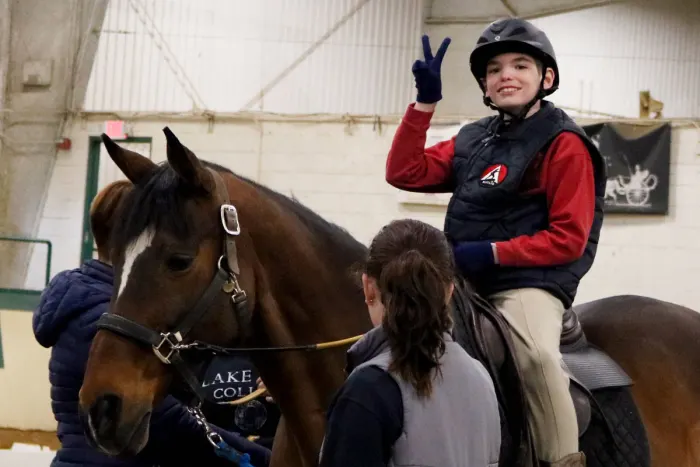 young female rider in a red shirt making a peace sign while riding a horse