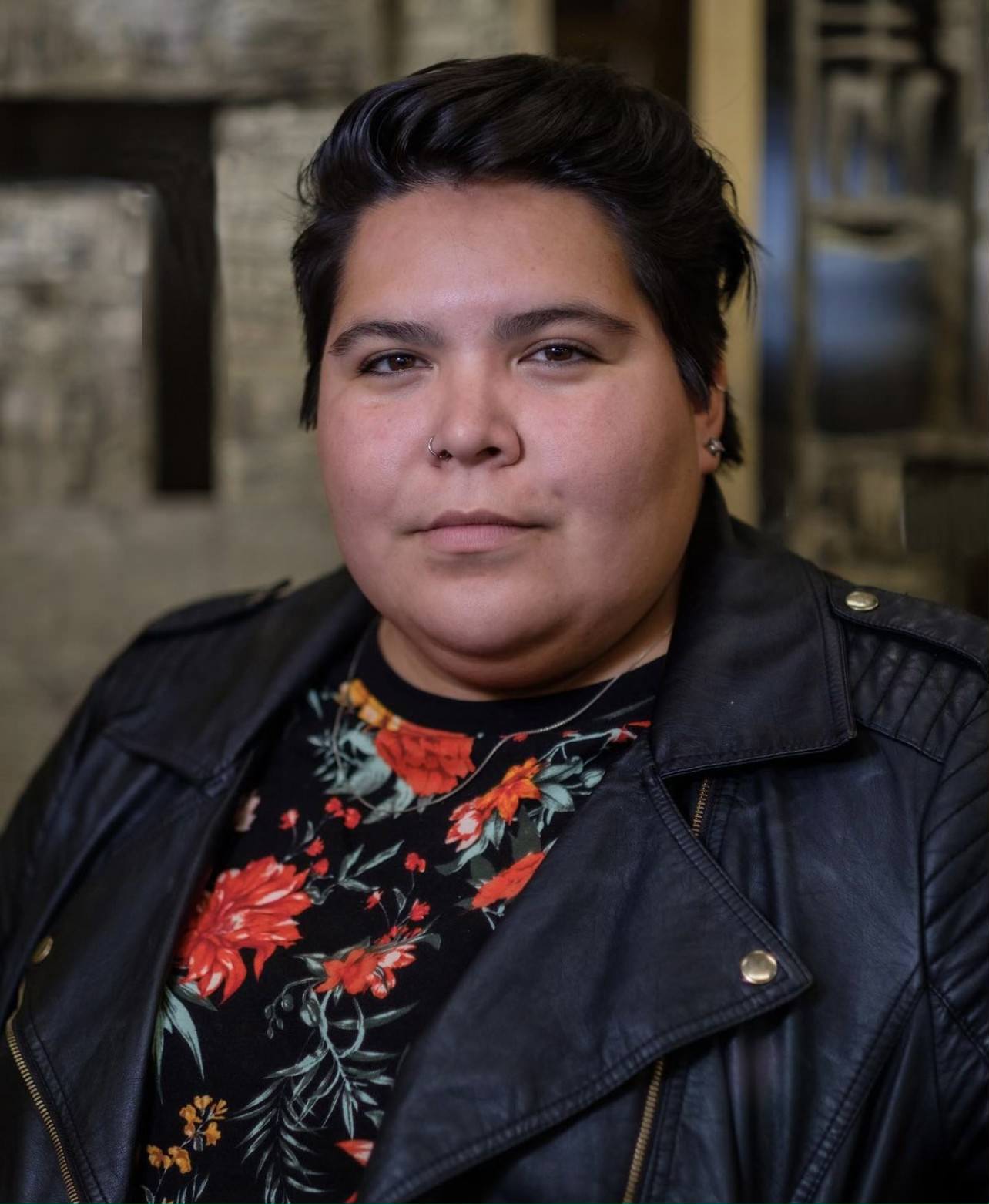Headshot of a woman with short hair combed backwards, leather jacket, and a shirt with flowers underneath. She looks at the camera