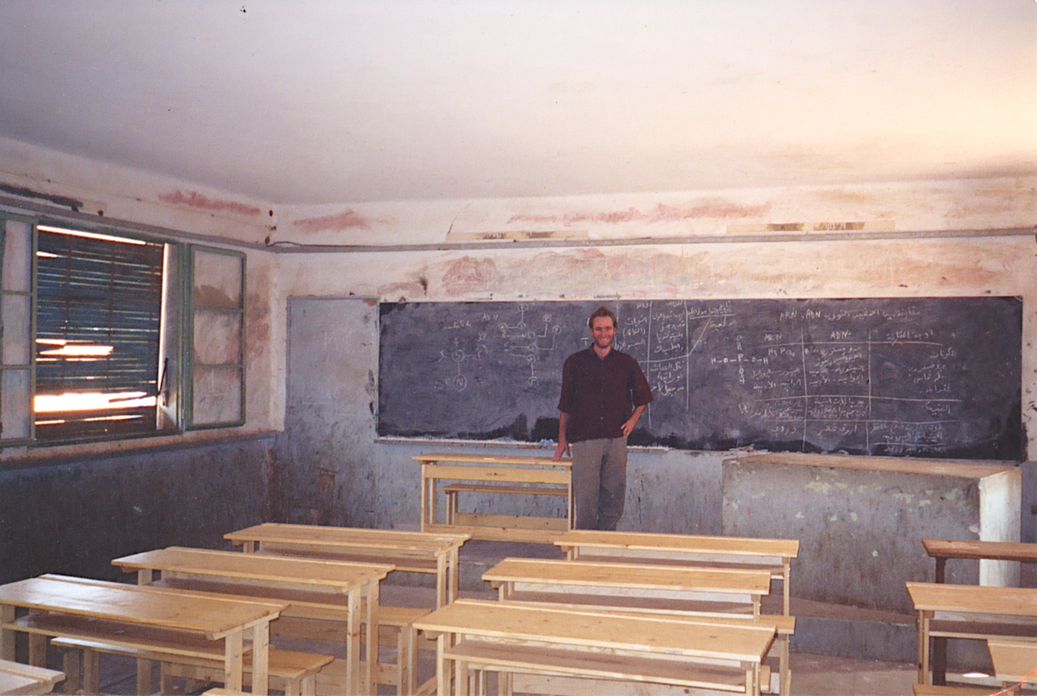 John is standing at the head of a classroom facing the viewer. Behind him is a dusty chalkboard nearly the length of the wall. In front of him stand several bench-style desks, for two kids each. The walls are dirty; paint is chipping off surfaces; the shutters in the window are blocking the sunlight except where they are broken.