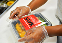 A close up look at gloved hands sealing a package that contains a Newark Working Kitchen meal. The NWK logo is prominent on the meal package's cover.