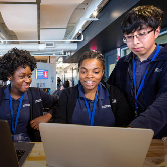 Audible's high school interns stand behind a table in our cafeteria at our offices in Newark. Two laptops sit open in front of them and they are looking at one of them as if conferring about something open on it.