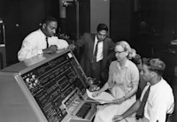 Several individuals working on and around a large, vintage computer console with numerous dials and switches, in a room filled with various electronic equipment.