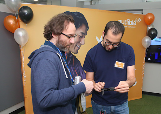 Two men stand close looking down at one of the men's phones. They are in front of a large orange poster displaying a man listening to Audible who appears, due to the angle of his head and that it falls between the two mens' shoulders, to be peering at their phone too.