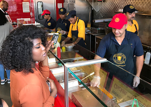 A woman in an orange shirt stands at a deli counter holding a Lunch Out Wednesday card. A restaurant employee in a red cap is serving her.
