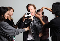 David Spade stands center with a polaroid camera in hand, snapping a photo, while four female stylists surround him prepping his hair and clothing for a photo shoot.