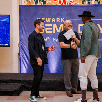 Two men accept a congratulations from another man in front of a Newark Demo Day backdrop.