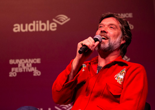 Rufus Wainwright speaks into a microphone at the Sundance event, "A Conversation with Rufus Wainwright." He's wearing a red button down and sitting in front of Audible signage.