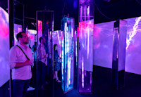 A man with glasses is standing in a dark art installation in which purple light is being reflected on mirrors placed throughout the small room.