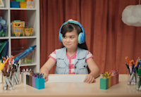 A photo of a young girl wearing large blue headphones. She’s seated at a desk which is covered in a colorful selection of Crayola markers, colored pencils and crayons.