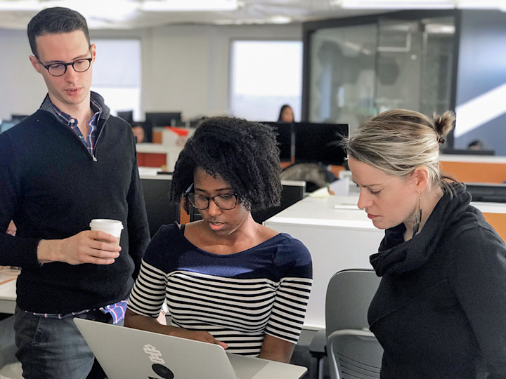 Two colleagues look over the shoulder of a woman working on a laptop computer.