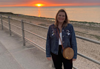 Audible employee Jess Falconer stands on a beach boardwalk. Behind her is a railing and then below it a sandy beach. The sun is setting over the ocean, a red burning glow in the distance. Jess is looking straight to the camera, smiling, while wearing a white t-shirt, jean jacket, black pants and a crossbody straw bag.