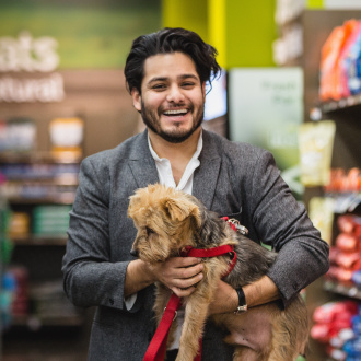 A man carries his small yorkie dog, who is wearing a red leash, through a lobby in Newark. He is smiling and wearing a white button down shirt and grey blazer.