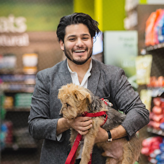 A man carries his small yorkie dog, who is wearing a red leash, through a lobby in Newark. He is smiling and wearing a white button down shirt and grey blazer.