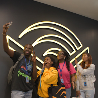 Four students stand in front of a neon Audible logo take a photo selfie.