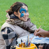 A girl sits in a chair having her face painted with a blue leave.