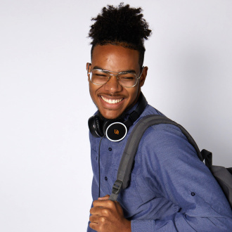 A teen stands sideways against a neutral background facing toward the camera with a big smile on his face. He has a blue button down shirt on, Audible headphones sitting around his next, and a grey backpack over his left shoulder. 