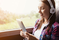 A girl sits by the window on a bus wearing a white t-shirt and a red and blue flannel shirt with silver headphones on. She's listening to a Audible book playig on the tablet she's holding.