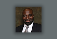 Audible employee St Jules Desir appears against a gray background wearing a blue suit, white button down shirt and pink and silver patterned tie. He is looking directly at the camera and smiling.